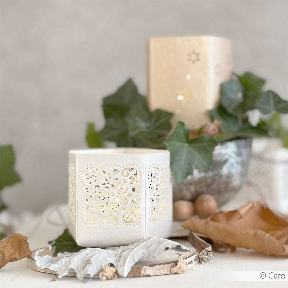 Weiße, dekorative Kerzenhalter von Alexandra Renke mit filigranen Mustern, die den Windlicht-Ornamenten von Motivstanze ähneln, sind von grünem Efeu und getrockneten Herbstblättern umgeben.