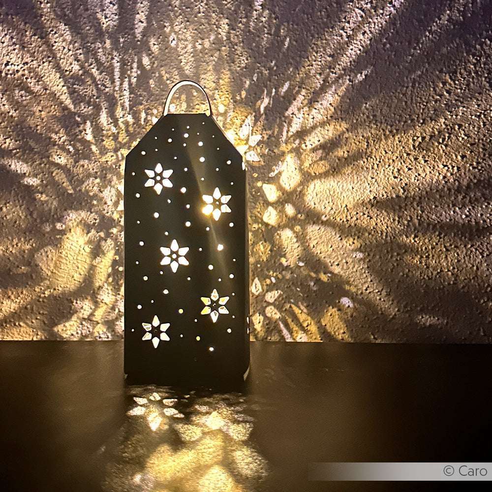 Eine Alexandra Renke Windlicht Ornament Laterne wirft sternförmige Lichtmuster auf strukturierte Wände und Oberflächen in einem dunklen Raum.