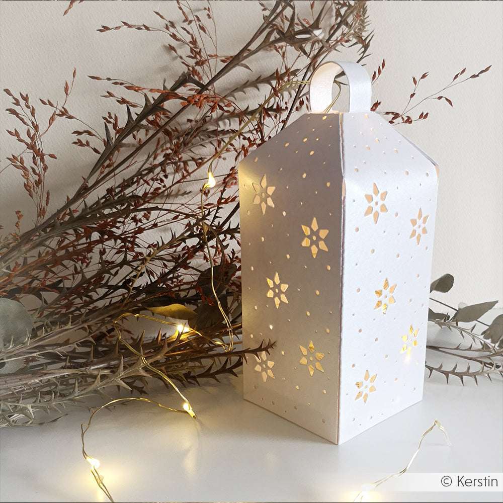 Weiße Papierlaterne mit ausgeschnittenen Schneeflocken, die mit Lichtern leuchten und von Lichterketten umgeben sind. Gebastelt mit Alexandra Renke Deep Edge Motivstanzen und PAPPcut®ies für einen detaillierten Windlicht-Ornament-Effekt.