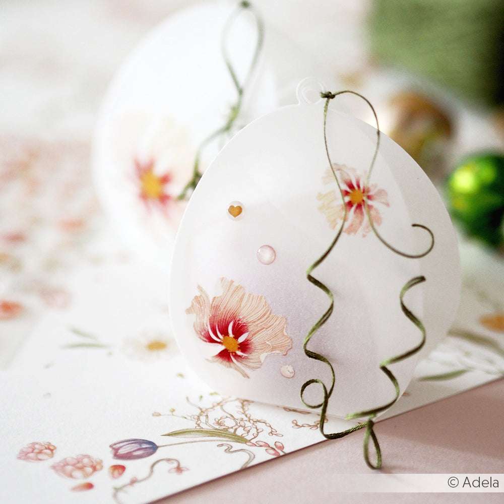 Zartes weißes Ornament mit rosafarbenen Blumenmustern und grünem Band auf einer floral gemusterten Oberfläche, perfekt zum Verzieren einer Alexandra Renke PAPPcuties Motivstanze Osterei Box.