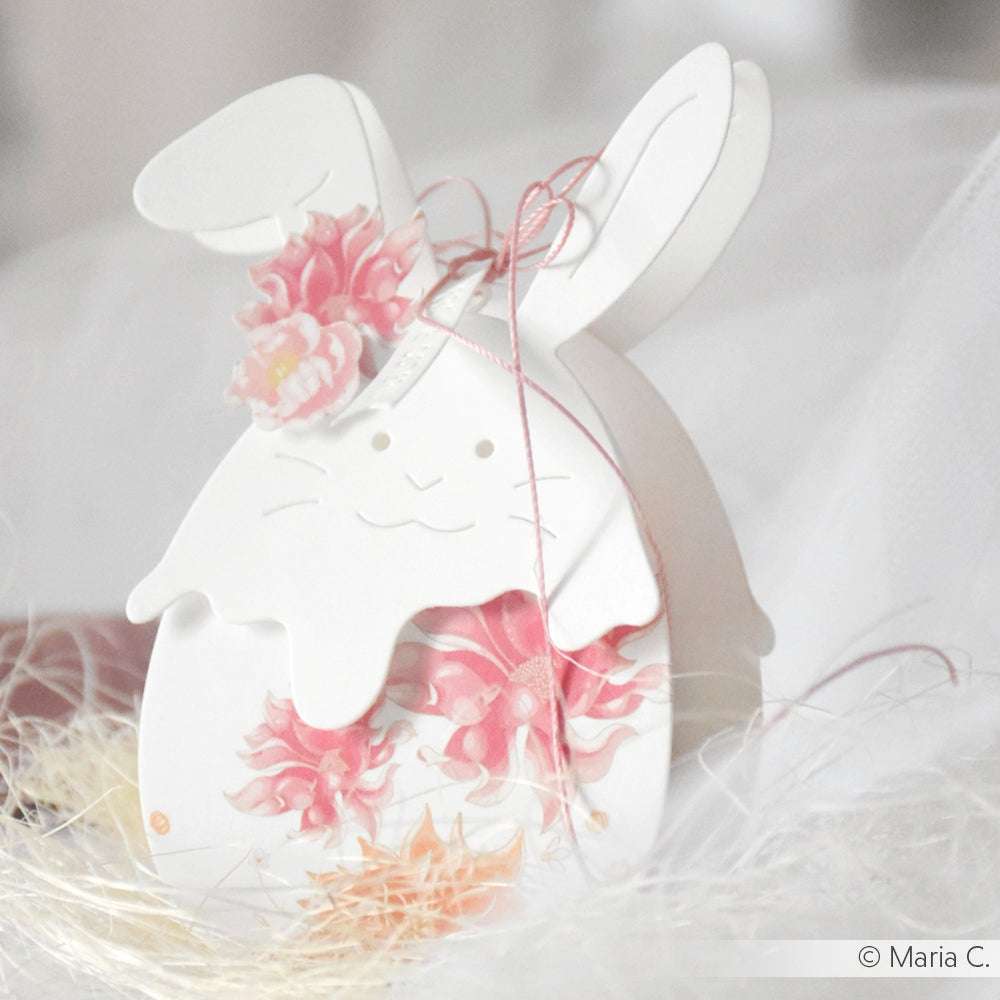 Eine weiße Karte in Hasenform von Alexandra Renke, hergestellt mit PAPPcut®ies und verziert mit rosa Blumen und Kordel, vor einem weichen, hellen Hintergrund.