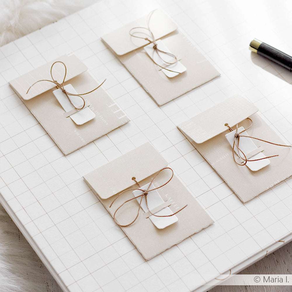 Vier cremefarbene, mit brauner Kordel gebundene Briefumschläge mit weißem Anhänger liegen auf einem Raster - fachmännisch gefertigt mit der Alexandra Renke Motivstanze Mini Umschlag für das besondere PAPPcut®ies Finish.