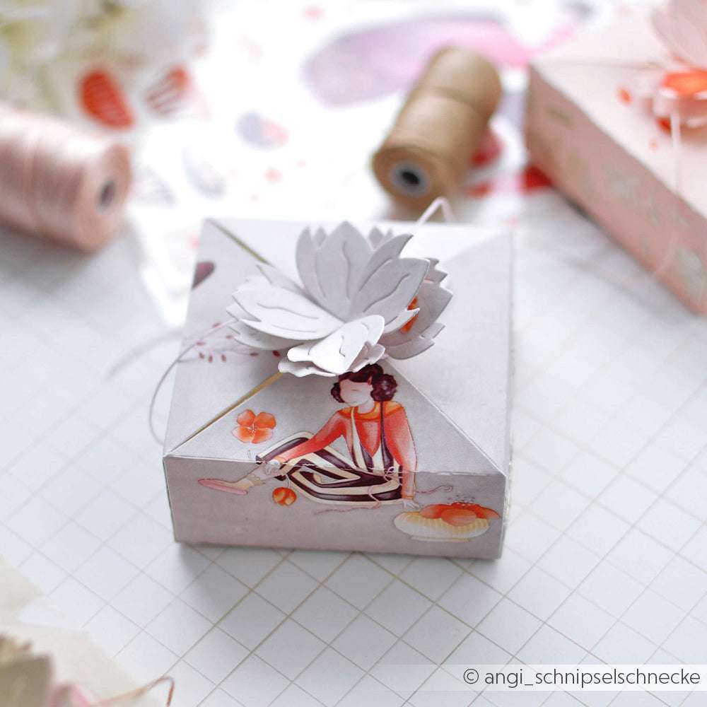Die dekorative Geschenkbox von Alexandra Renke zeigt einen floralen Papiertopper und die Illustration einer sitzenden Frau, umgeben von Bastelmaterial wie PAPPcut®ies und Deep Edge Motivstanzen.