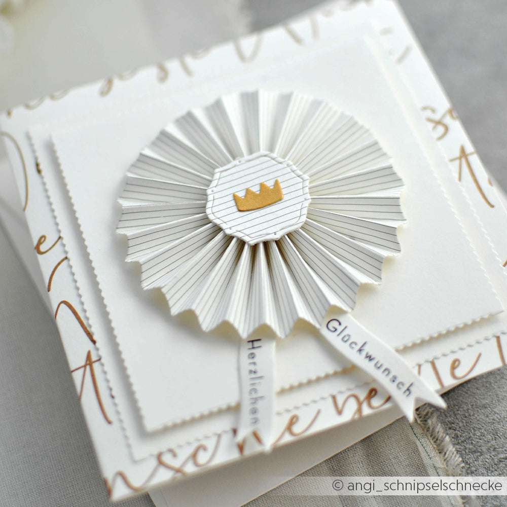 Eine weiße, gefaltete Papierrosette, hergestellt mit der Alexandra Renke PAPPcut Motivstanze Rosette, verziert mit einer goldenen Krone und deutschem Text auf einer dekorativen Karte.