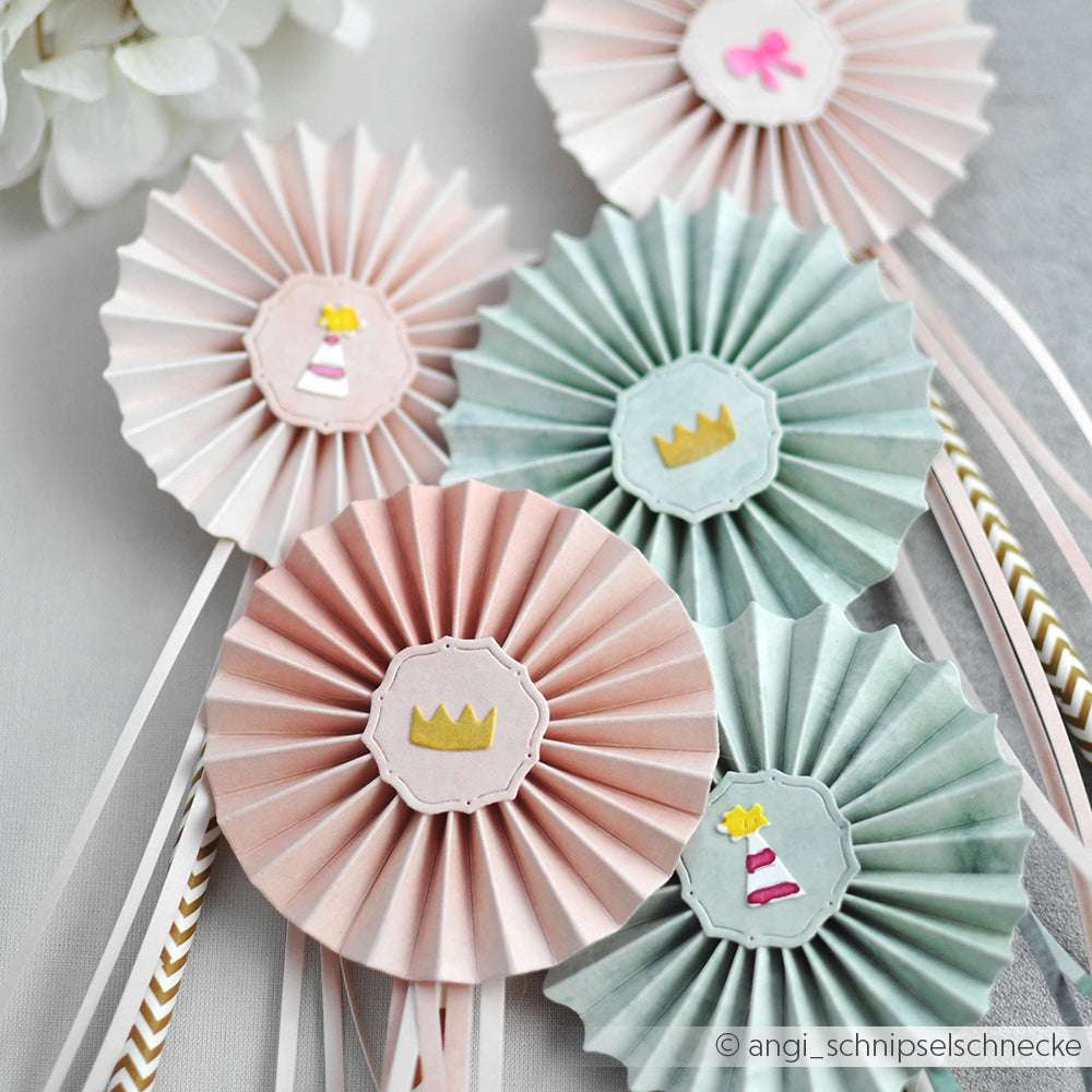 Fünf pastellfarbene Papierrosetten von Alexandra Renke, hergestellt mit PAPPcut®ies und Deep Edge Motivstanzen, jeweils verziert mit Kronen, Partyhüten und Bändern für einen festlichen Akzent auf einer hellen Fläche.