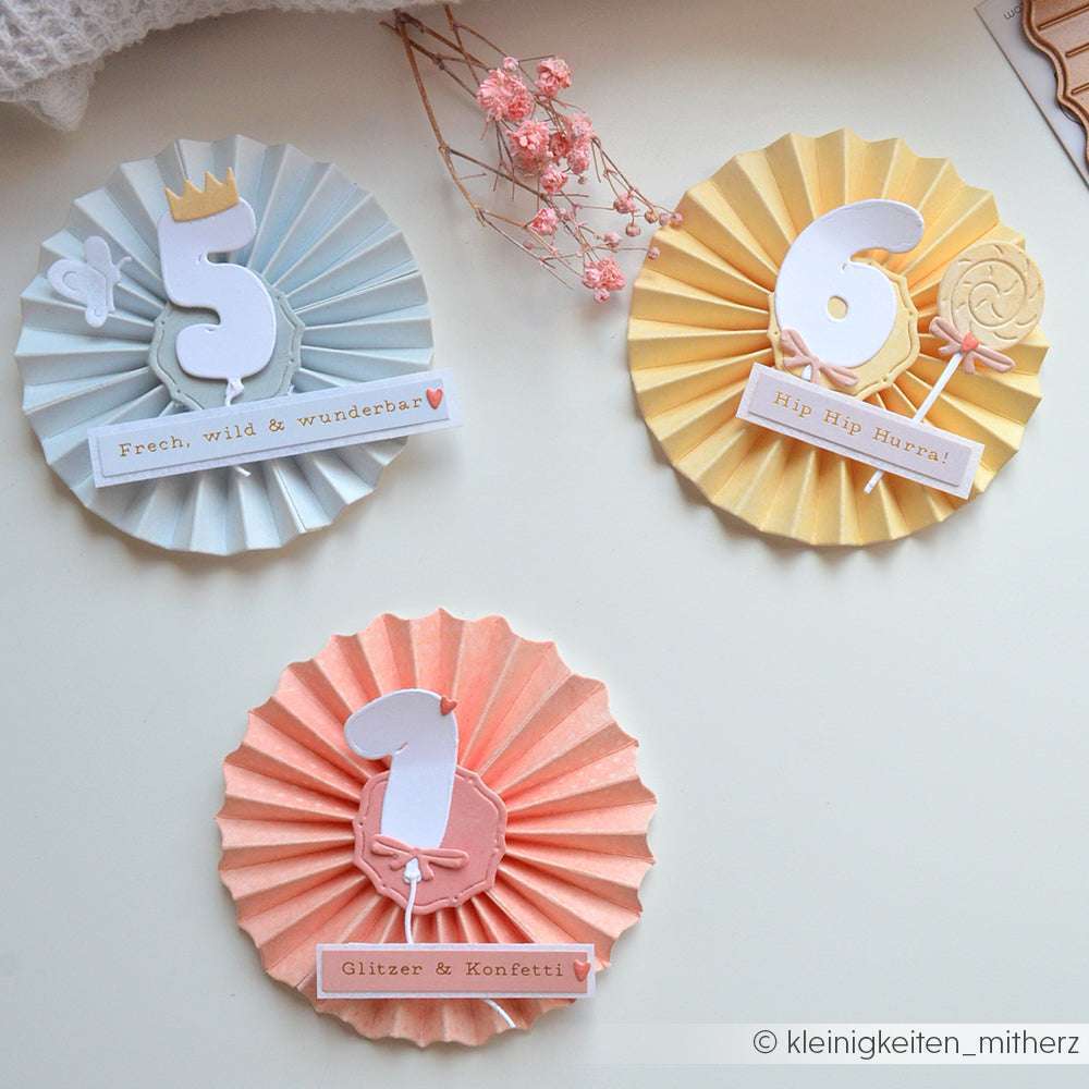 Drei farbenfrohe Papierrosetten mit den Zahlen 5, 6 und 1, die jeweils mit der Alexandra Renke Motivstanze Rosette ausgestanzt wurden, perfekt für eine festliche Geburtstagsfeier.