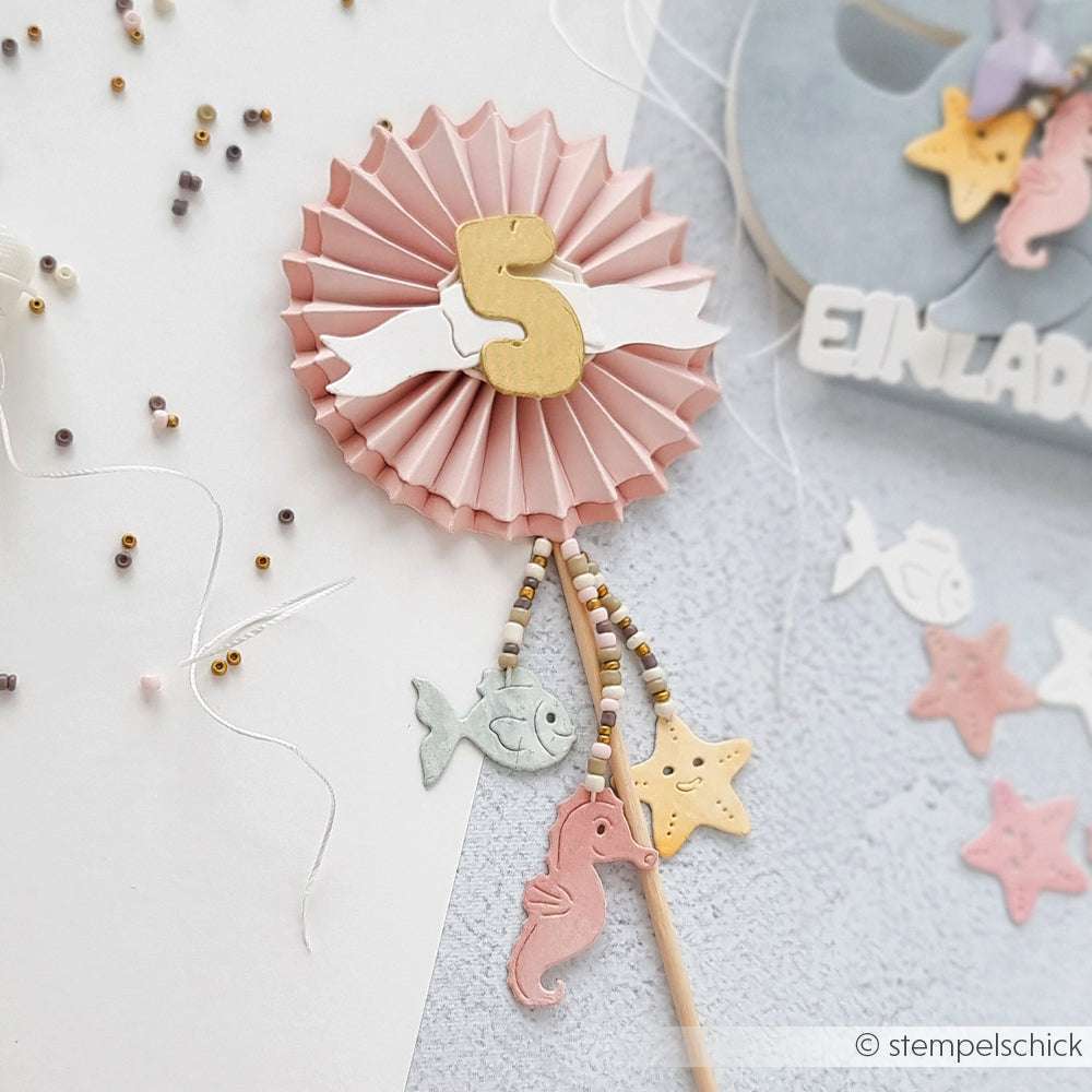 Rosafarbene Rosette mit einer goldenen Nummer 5, hängenden Fischen, einem Seepferdchen und Sternen, die mit Alexandra Renke Deep Edge Motivstanzen und PAPPcut®ies hergestellt wurden, verziert mit Perlenschnüren für einen charmanten Touch.