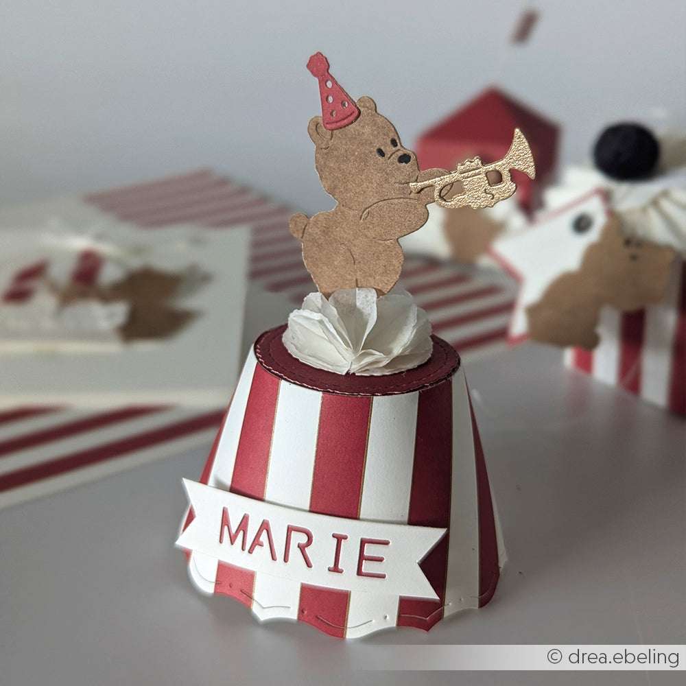 Die bezaubernde Alexandra Renke Papierbastelei eines Bären mit Partyhut, der Trompete spielt, auf einem gestreiften Sockel mit der Aufschrift MARIE. Perfekte Cupcake- oder Muffin-Banderole PAPPcut®ies - macht jede Feier zu etwas ganz Besonderem!.