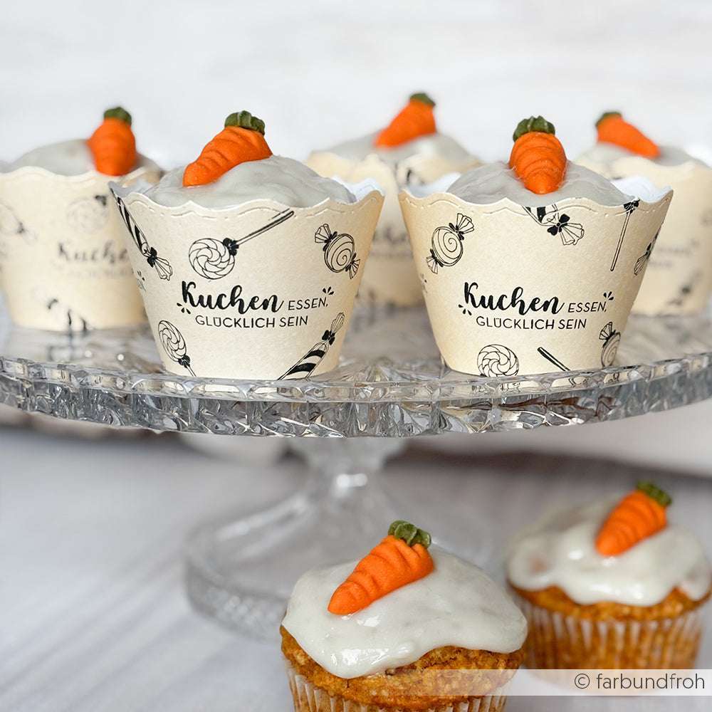 Alexandra Renke Karotten-Cupcakes mit weißem Zuckerguss und Fondant-Karotten, jeweils in einer stilvollen Muffin-Banderole, elegant präsentiert auf einem Tortenständer aus Glas.