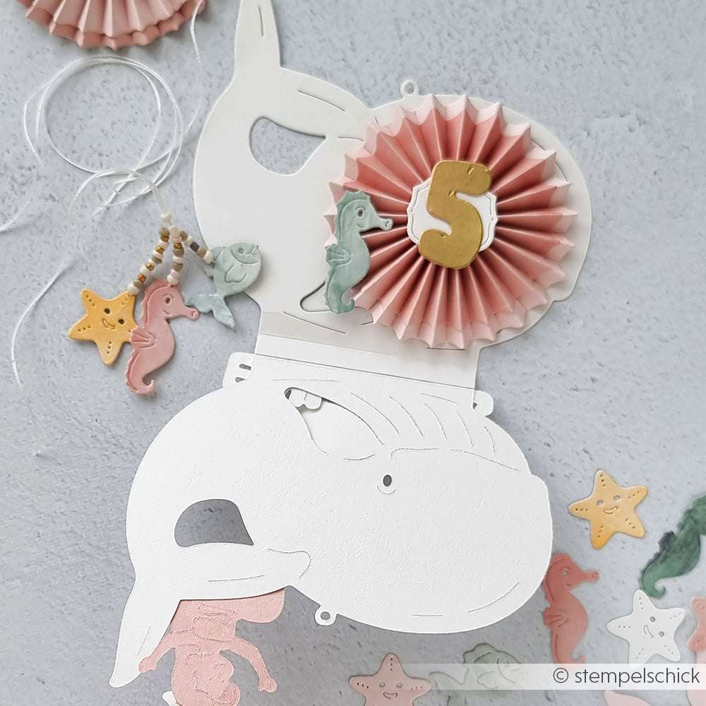 Weißer, walförmiger Papierausschnitt von Alexandra Renke, verziert mit einer rosafarbenen Rosette, der goldenen Nummer 5, Seepferdchen-Anhängern, Seestern-Akzenten und feinen Motivstanze-Details aus der PAPPcut®ies Kollektion.