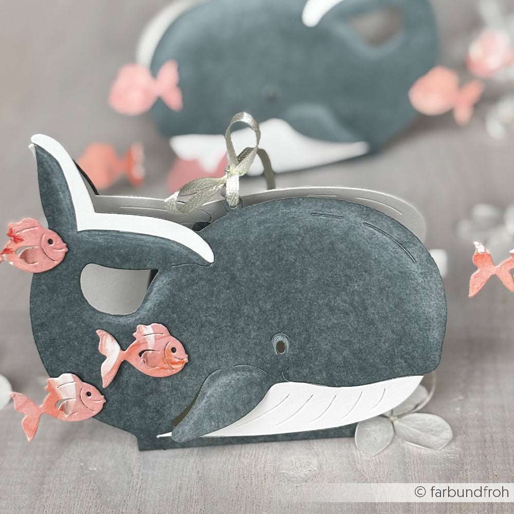 Eine graue walförmige Geschenkbox, gefertigt mit der Alexandra Renke PAPPcut®ies Motivstanze Fröhliche Wal-Box und verziert mit kleinen rosa Fischen und einer silbernen Schleife, ruht elegant auf einer hellen Fläche.