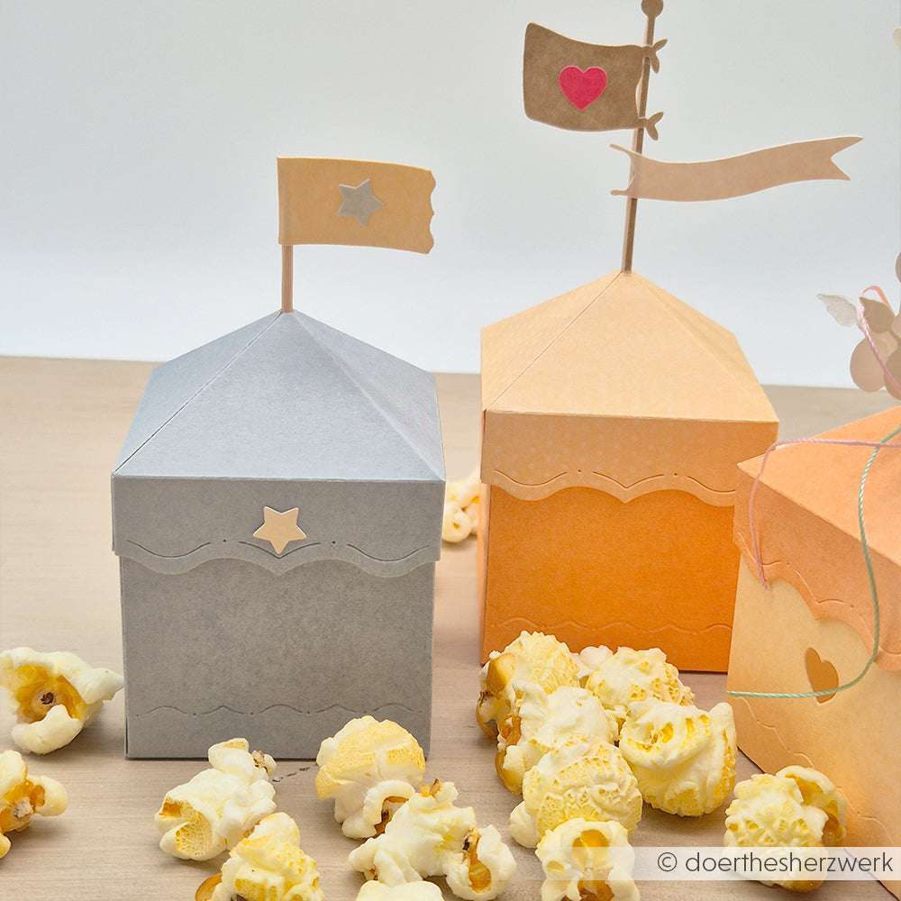 Zwei dekorative Alexandra Renke PAPPcuties Popcorndosen in Form von Zelten mit Fahnen, umgeben von Popcorn auf einer Holzoberfläche.