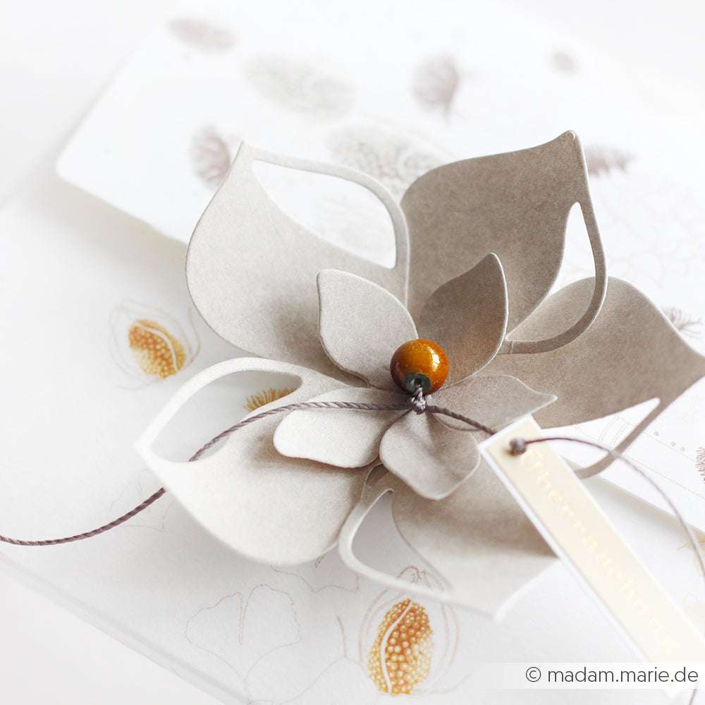 Eine zarte Papierblume mit Perlen in der Mitte und einem Anhänger, hergestellt mit Alexandra Renke Deep Edge Motivstanzen, ruht auf einem weißen Umschlag, der mit Blumenmustern verziert ist.