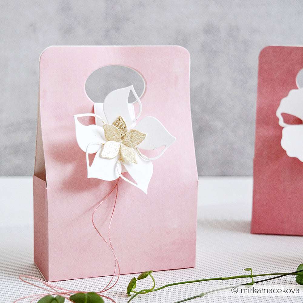 Rosafarbene Geschenktüte mit einer weiß-goldenen Blumendekoration aus der Alexandra Renke Motivstanze Faltzauber-Blume, die auf einer hellen Fläche platziert wird - perfekt, um jede Feier mit PAPPcut®ies zu verschönern.