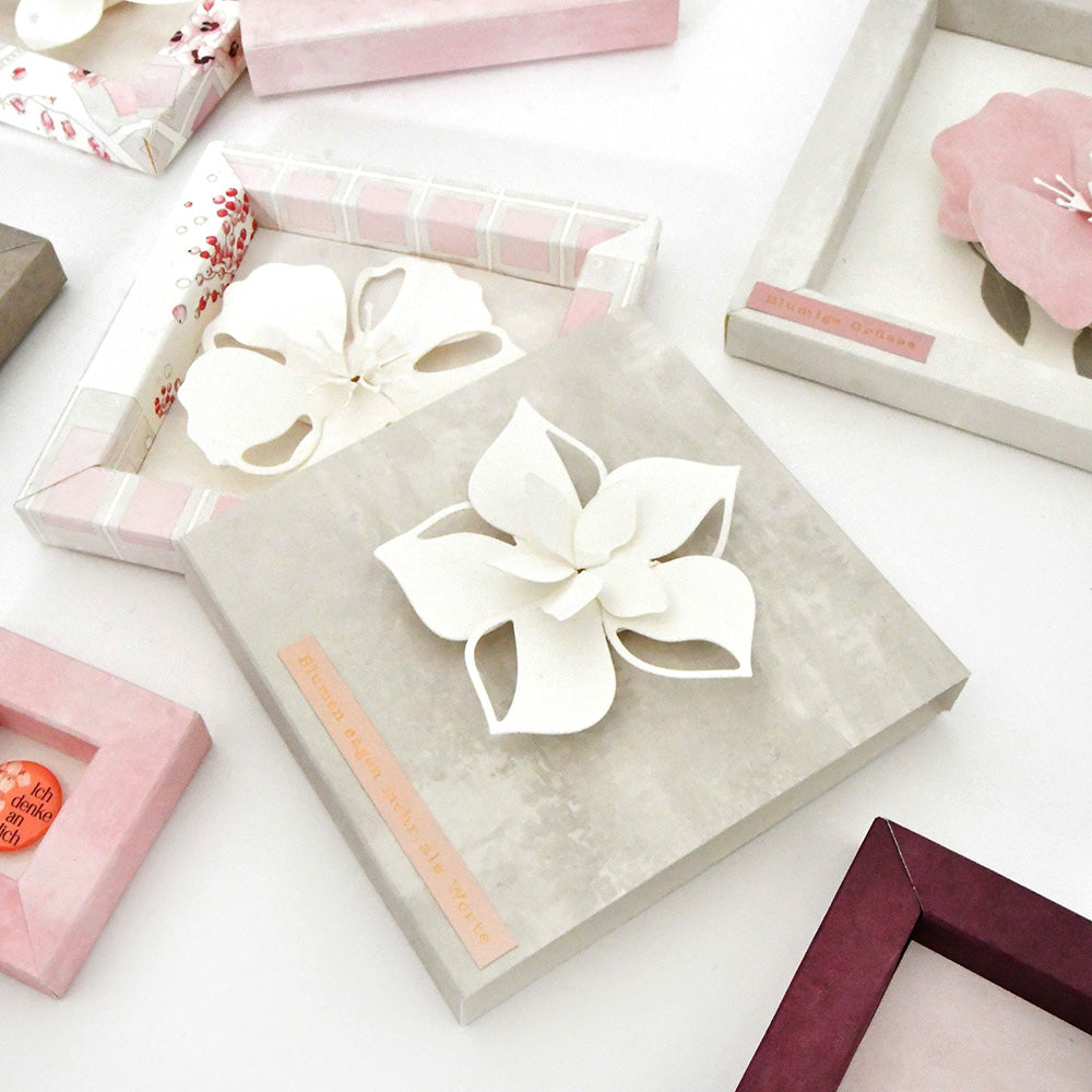 Schattenboxen mit Papierblumen aus Alexandra Renke Motivstanze Faltzauber-Blume und PAPPcut®ies, die in pastellfarbenen Rahmen auf einer weißen Fläche präsentiert werden.