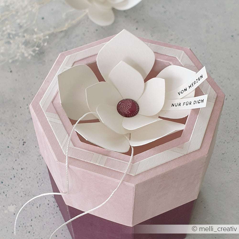 Eine rosafarbene sechseckige Geschenkschachtel, verziert mit einer weißen Papierblume, die mit der Motivstanze Faltzauber-Blume 3 von Alexandra Renke gestaltet wurde, und Anhängern mit der Aufschrift VON HERZEN NUR FÜR DICH.