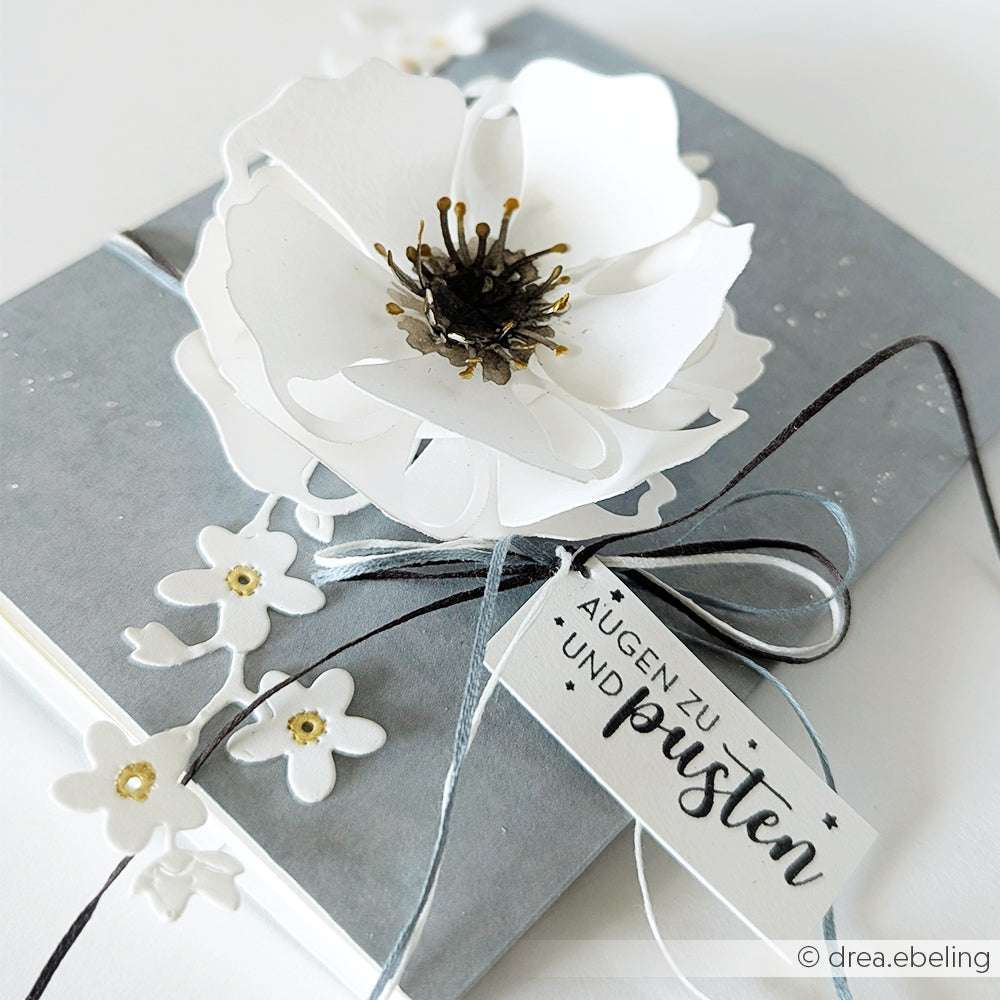 Das Geschenk ist in grauem Papier mit einer weißen Blume, schwarzer Schnur und einem Anhänger mit der Aufschrift "AUGEN ZU UND pusten" verpackt und mit einem eleganten PAPPcut-Detail von Alexandra Renke veredelt.