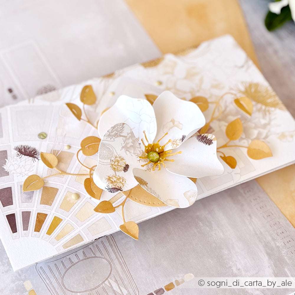 Eine handgefertigte Karte zeigt eine große weiße Papierblume, die mit der Alexandra Renke Motivstanze Faltzauber-Mohn 2 gestaltet wurde, akzentuiert durch goldene Blätter auf einem dekorativen Hintergrund.