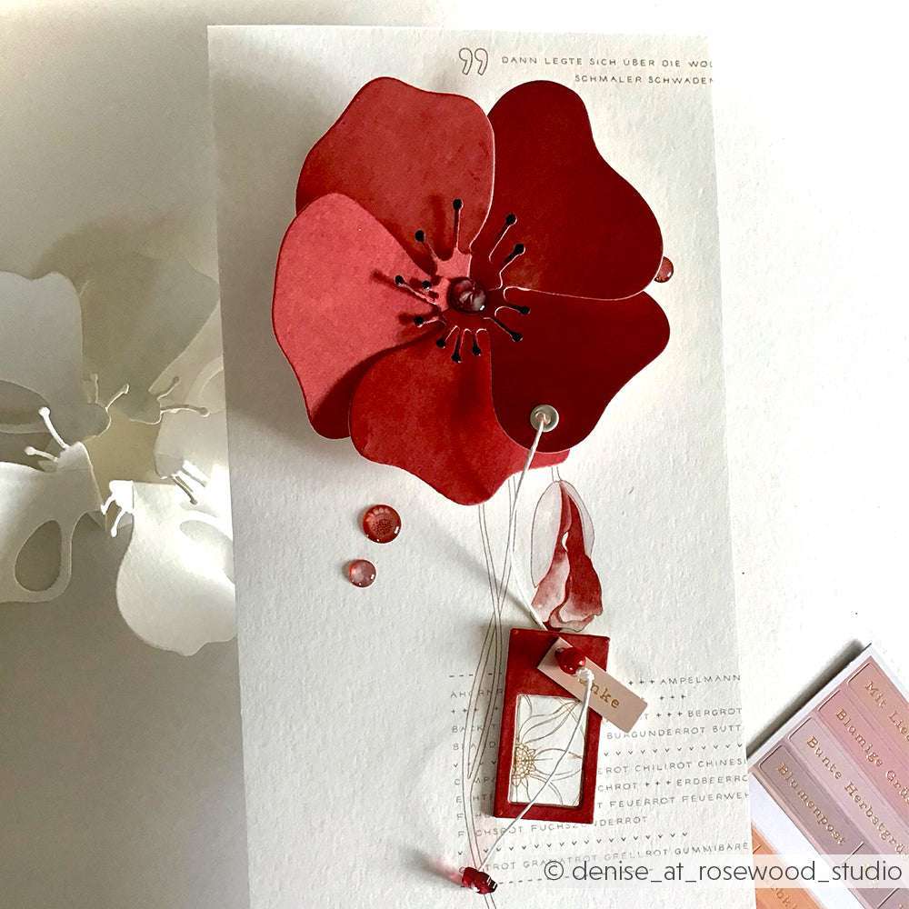 Handgefertigte Karte von Alexandra Renke mit einer großen roten 3D-Blume aus Faltzauber-Mohn 2 und dekorativen Anhängern auf weißem, strukturiertem Hintergrund.