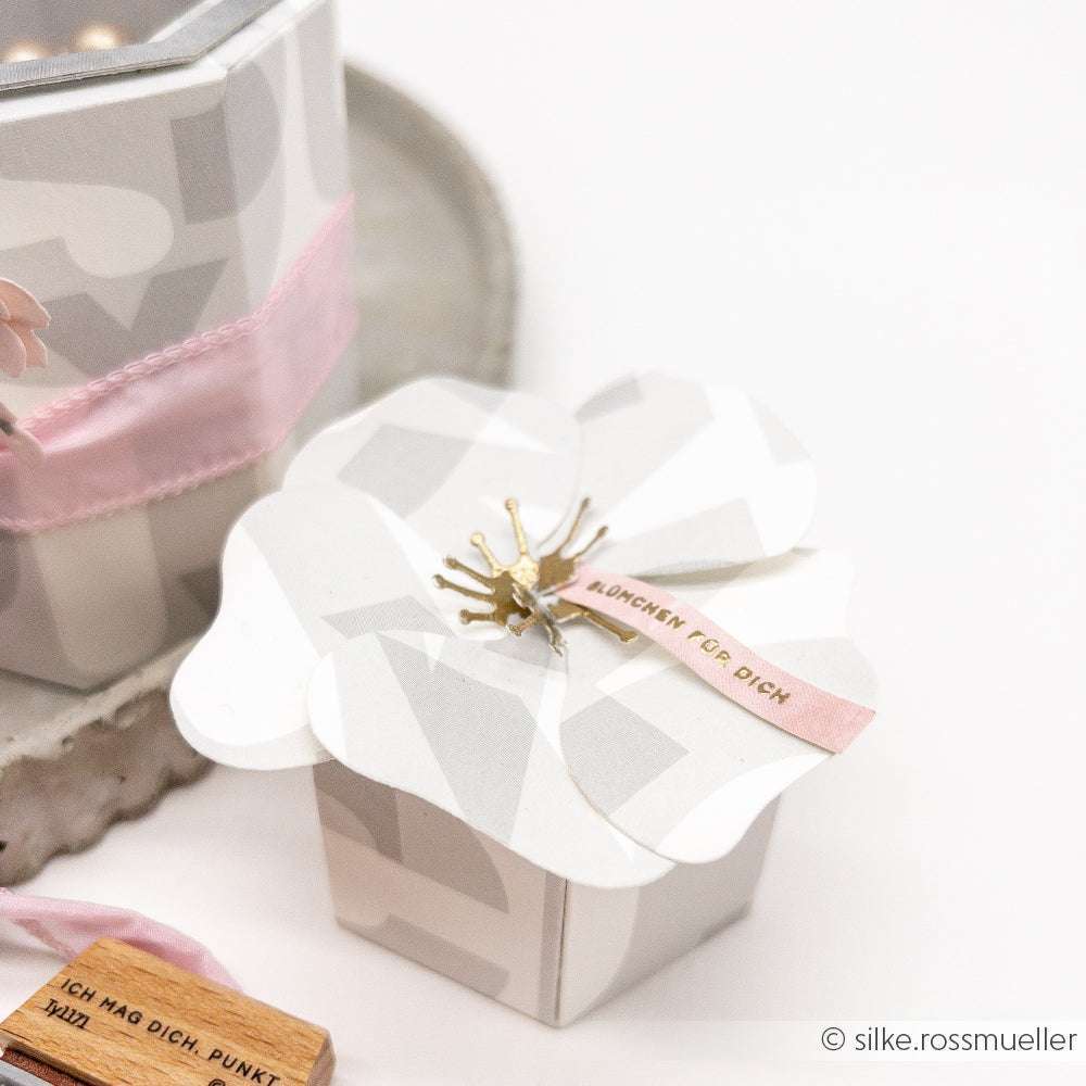 Weiße blumenförmige Geschenkschachtel mit goldener Mitte und rosafarbenem Bandanhänger, hergestellt mit Alexandra Renke Motivstanze Faltzauber-Mohn 2, neben einem Tablett und einer weiteren Schachtel.
