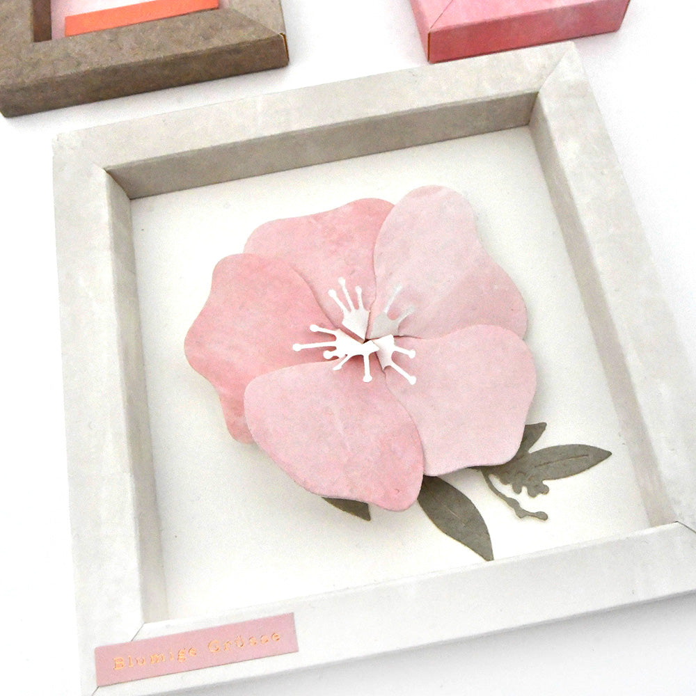 Eine gerahmte rosa 3D-Papierblume mit grünen Blättern, die mit Alexandra Renke Deep Edge Stanzen und PAPPcut®ies hergestellt wurde, steht auf einem sauberen weißen Hintergrund.