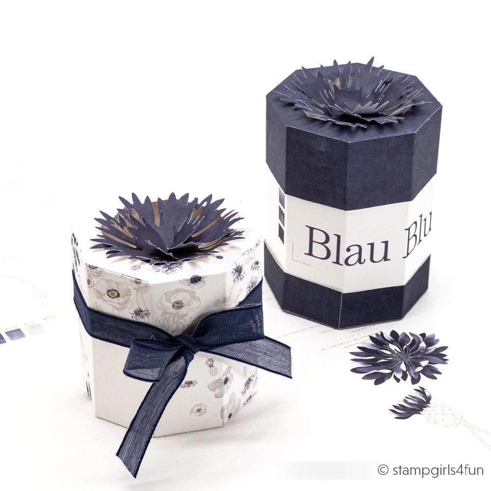 Zwei sechseckige Geschenkschachteln von Alexandra Renke, die mit dunklen Papierblumen und einem marineblauen Band verziert sind, zeigen elegante Motive aus PAPPcut®ies und der Motivstanze Faltzauber-Kornblume, arrangiert auf einer weißen Fläche.
