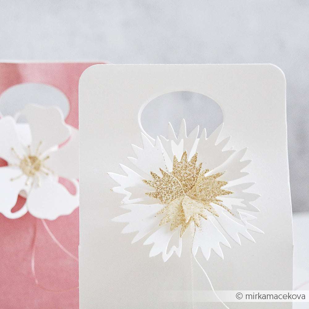 Weiße und rosafarbene Geschenktüten, verziert mit verschlungenen Papierblumen, die mit Alexandra Renke Deep Edge Punches und PAPPcut®ies Faltzauber-Kornblume hergestellt wurden, mit glitzernden goldenen Zentren.