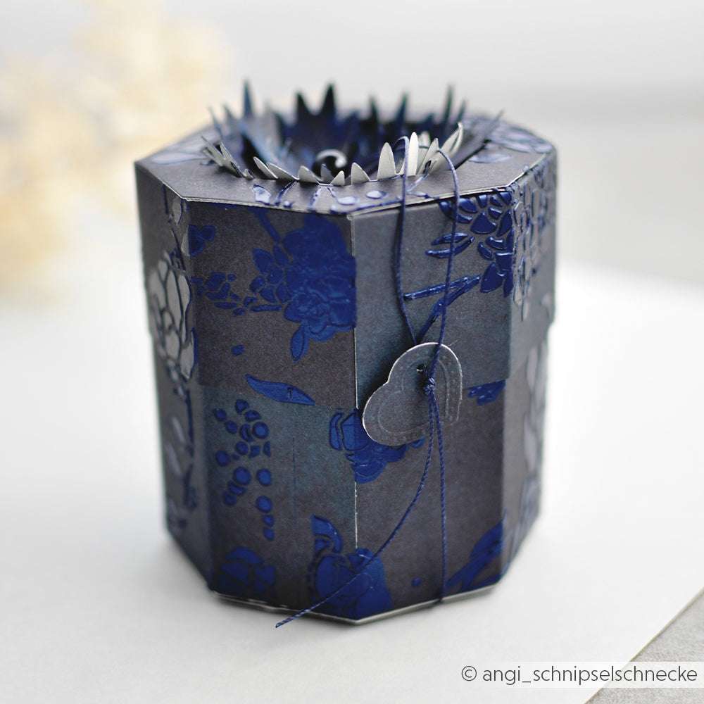 Eine achteckige Geschenkschachtel von Alexandra Renke in Dunkelblau mit floralem Muster, hergestellt mit Deep Edge Motivstanzen, mit einem dekorativen Deckel und einem herzförmigen Anhänger.