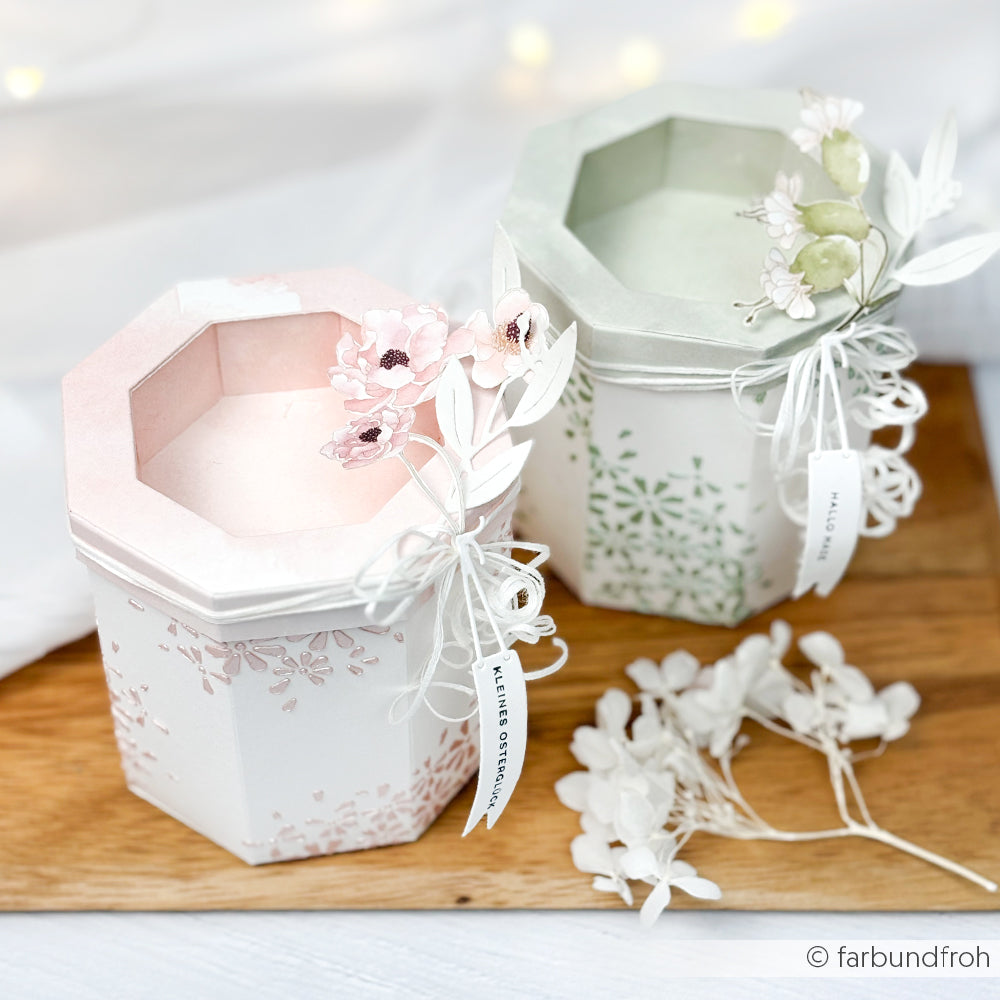 Zwei dekorative Alexandra Renke Achteckboxen mit Fenster, die mit pastellfarbenen Blumenmustern und Blumenverzierungen versehen sind, ruhen auf einer weißen Oberfläche - perfekt, um besondere Leckereien zu präsentieren.