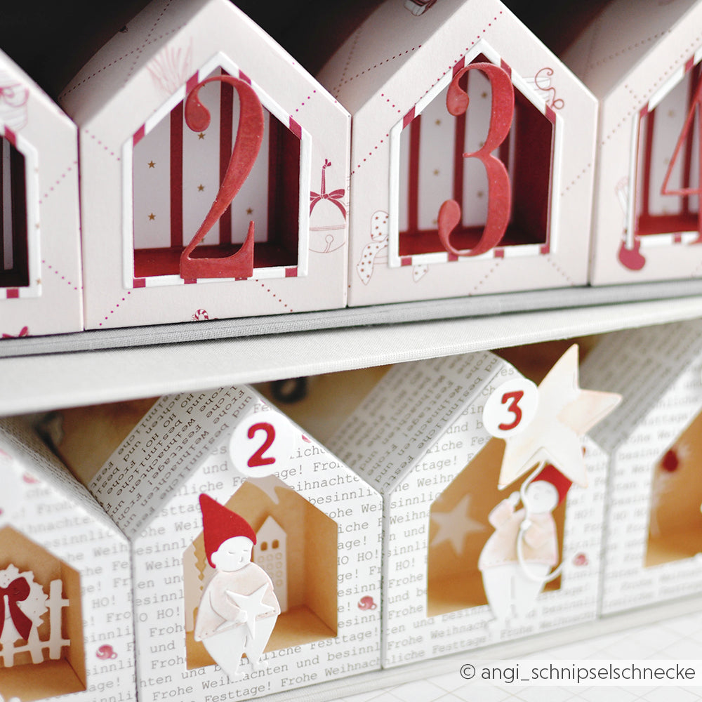 Eine Nahaufnahme eines Alexandra Renke Adventskalenders mit nummerierten, hausförmigen Fächern, gefertigt aus Designpapieren und veredelt mit einer Motivstanze Haus Box für präzise gestanzte Details, die zusammen mit festlichen Figuren präsentiert werden.