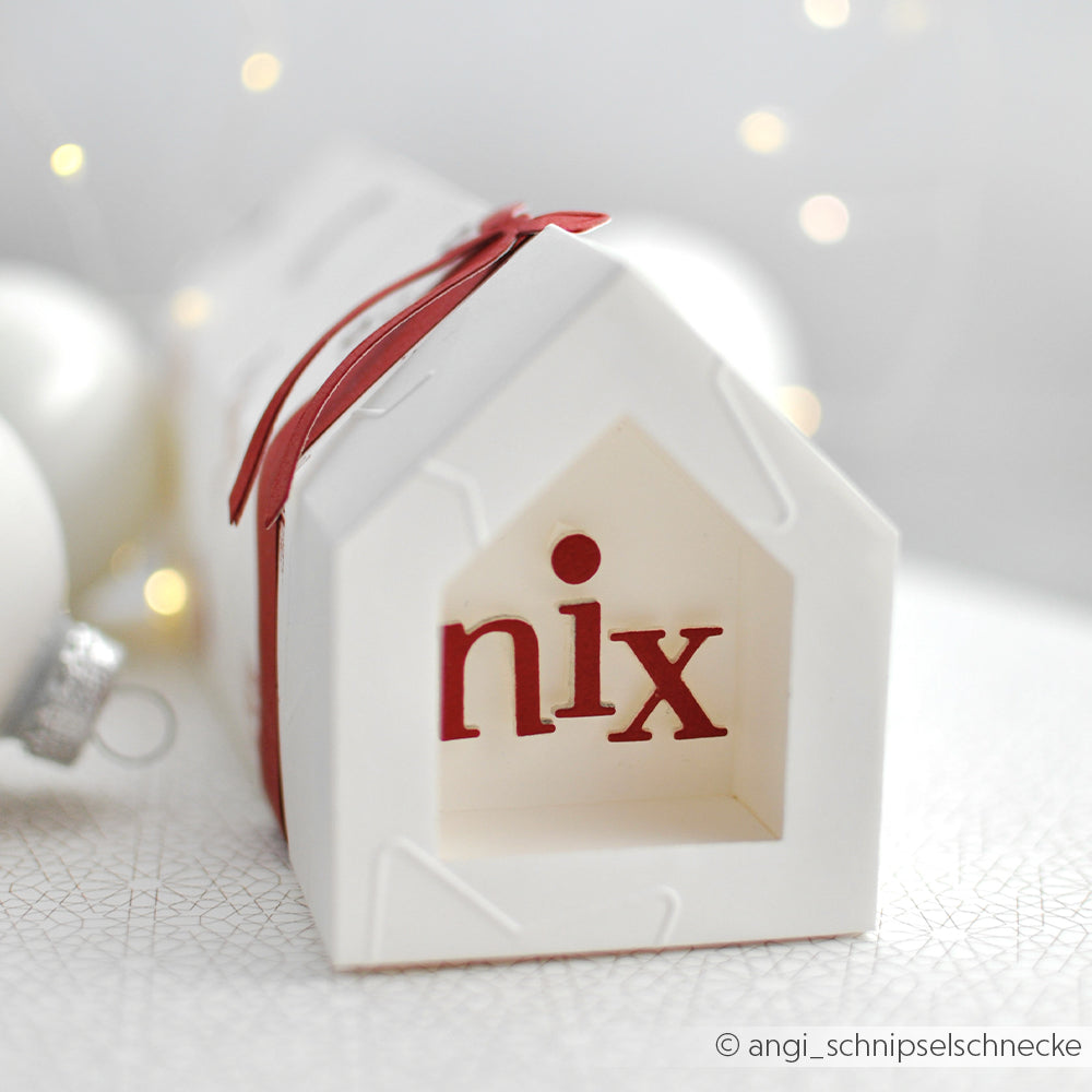 Auf einer weißen, hausförmigen Schachtel von Alexandra Renke, die mit der Motivstanze Haus Box gestanzt wurde, steht "nix" in roten Buchstaben und einem roten Band, daneben elegante weiße Ornamente.