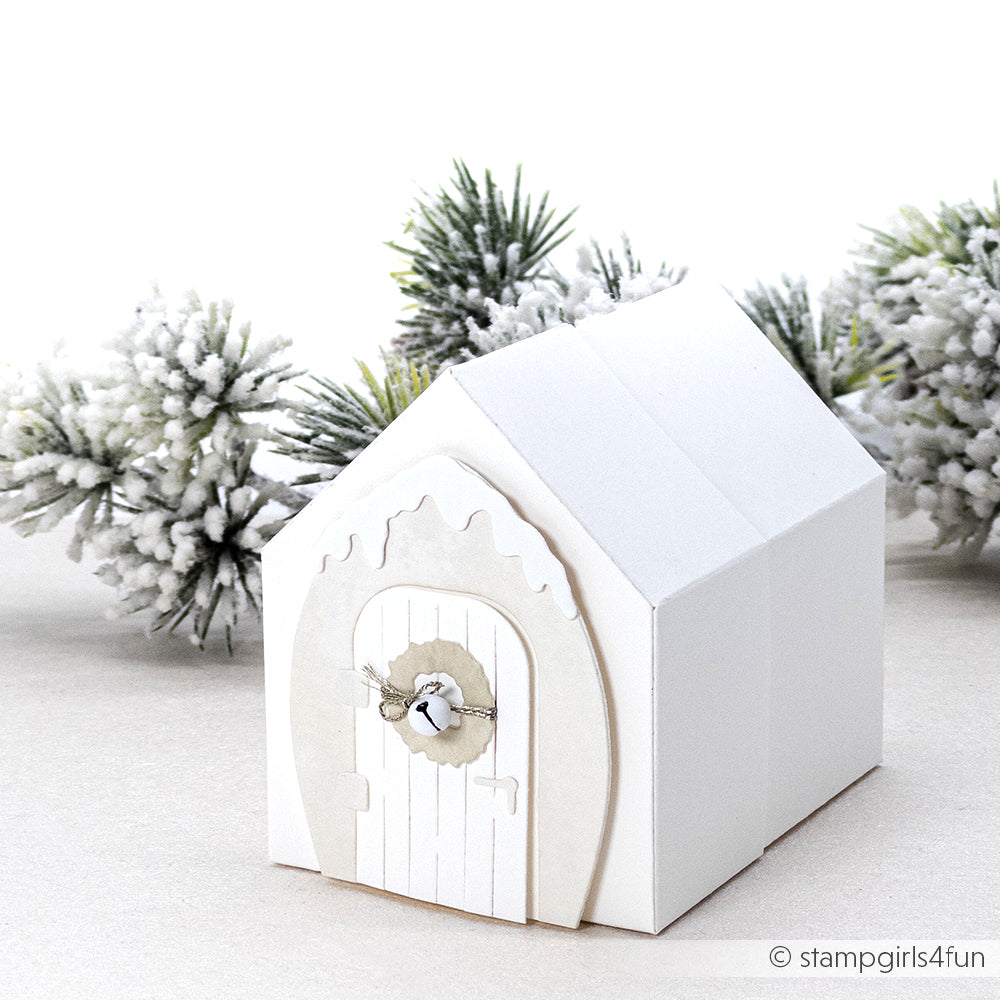 Vor verschneiten Tannenzweigen steht eine weiße Papierhaus-Geschenkbox mit runder Tür, gebastelt mit der Alexandra Renke Motivstanze Haus Box.