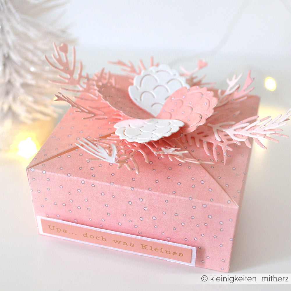 Eine rosafarbene, gepunktete Geschenkbox aus Alexandra Renke Designpapieren, verziert mit Papierblumen und Blättern, steht neben einem weißen Baum und Lichtern - perfekt, um Alexandra Renke Motivstanze Tannenzweig oder DieCut Systeme Projekte zu präsentieren.