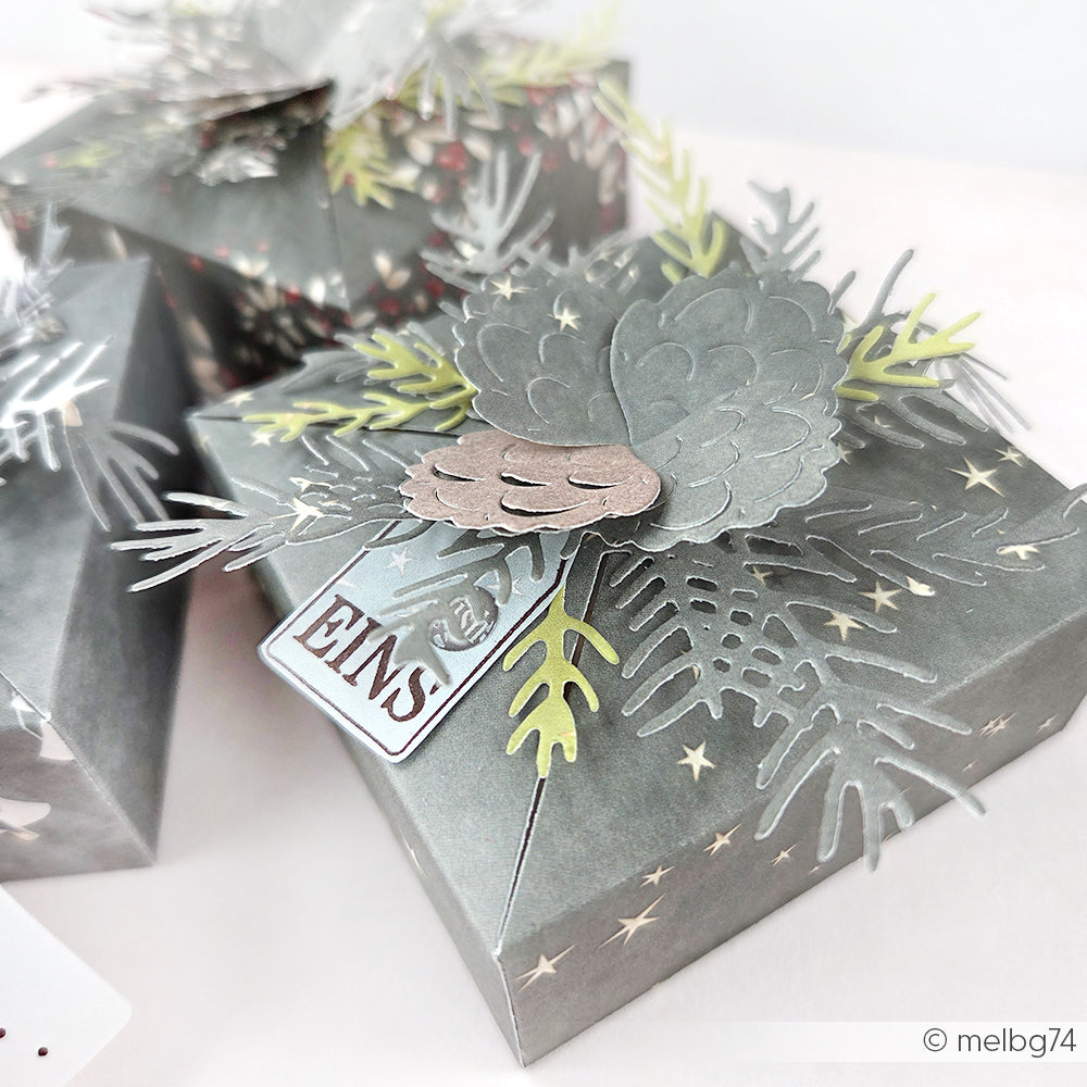 Graue Geschenkschachtel mit Sternenmustern, verziert mit Tannenzapfen und Blättern aus Papier, die mit der Alexandra Renke Motivstanze Tannenzweig ausgestanzt wurden, verziert mit einem kleinen Anhänger mit der Aufschrift ENS.