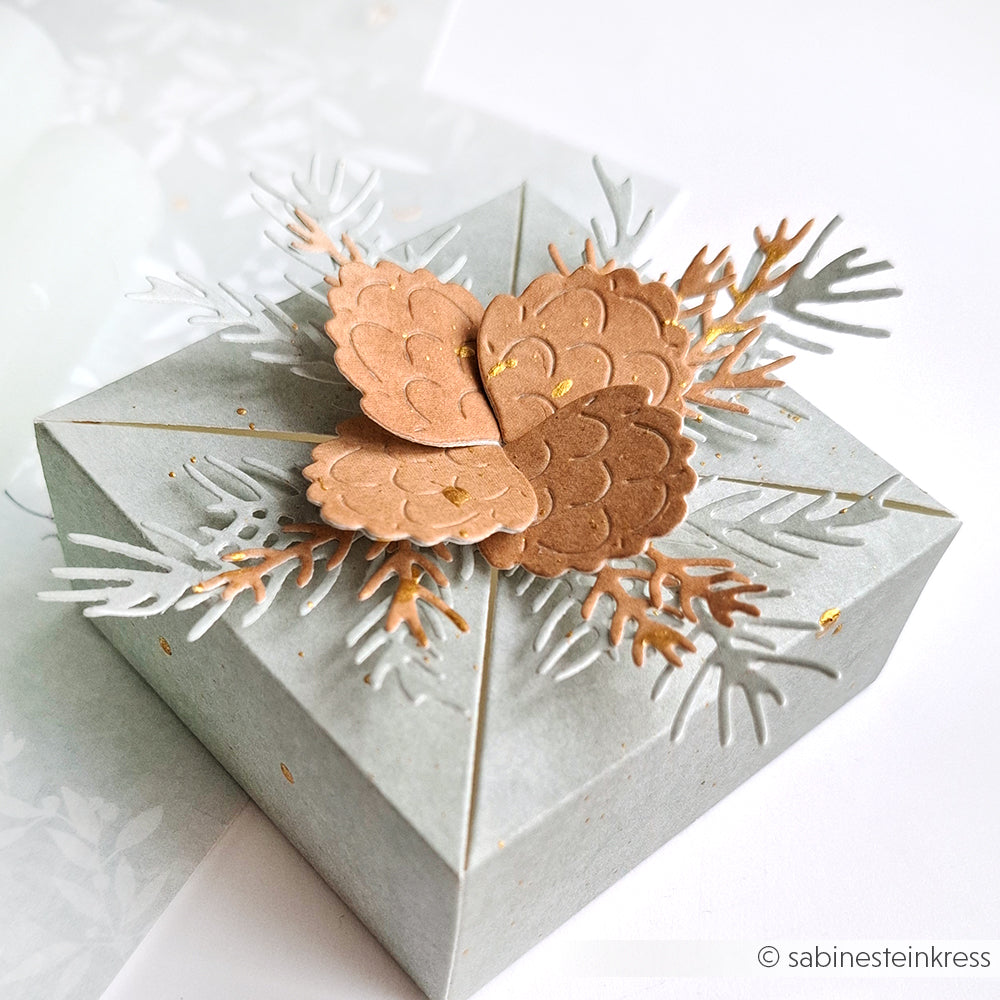 Ein Geschenkkarton mit Tannenzapfen aus Papier und weißem Laub auf dem Deckel, hergestellt mit Alexandra Renke Motivstanze Tannenzweig und Designpapieren 200g für eine festliche Note.