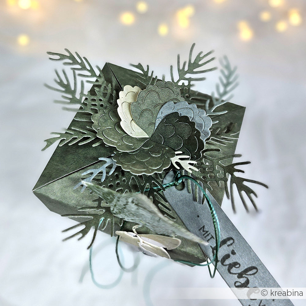 Ein grüner Geschenkkarton, verziert mit Tannenzapfen, Blättern und einem Bandanhänger, hergestellt mit Alexandra Renke Motivstanze Tannenzweig und eleganten Designpapieren, vor einem sanft verschwommenen Hintergrund.