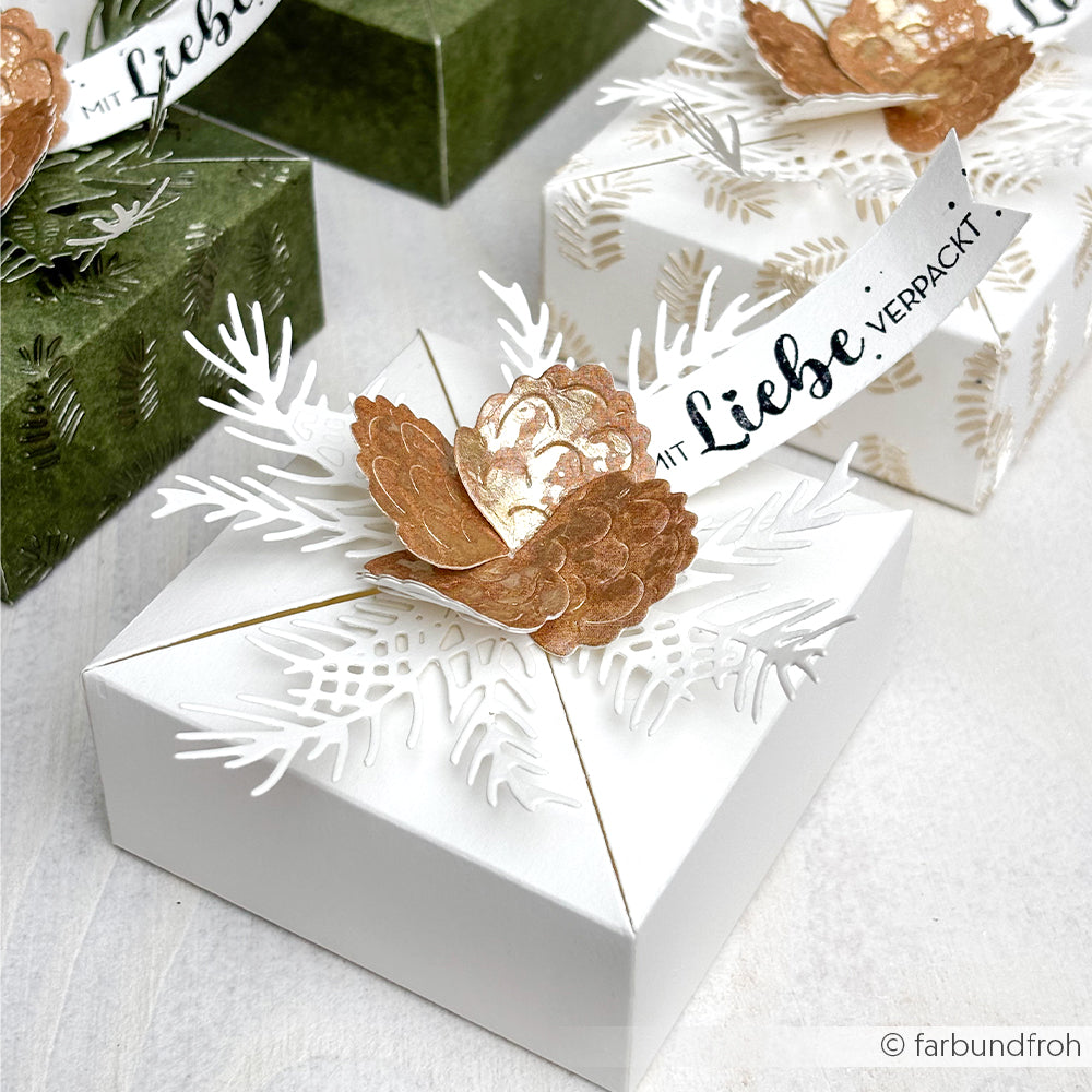 Weiße Geschenkschachtel mit goldener Blüte, weißem Laub, das mit Alexandra Renke Motivstanze Tannenzweig hergestellt wurde, und einem "Mit Liebe verpackt!"-Anhänger; im Hintergrund weitere Schachteln, die mit Alexandra Renke Designpapieren gestaltet wurden.
