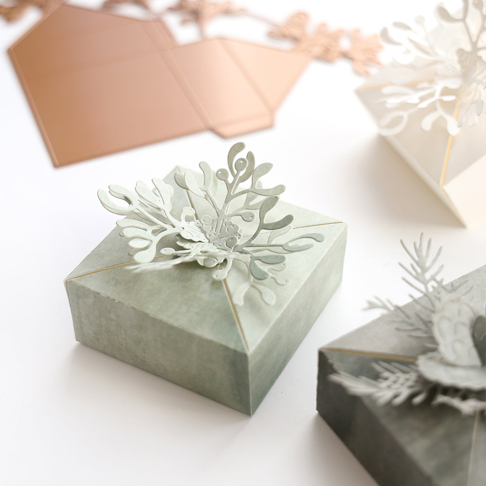 Obenauf liegt eine grüne Alexandra Renke Mistelzweig-Box mit komplizierten Blumenausschnitten, die mit einer Motivstanze hergestellt wurden, umgeben von anderen Bastelkisten.