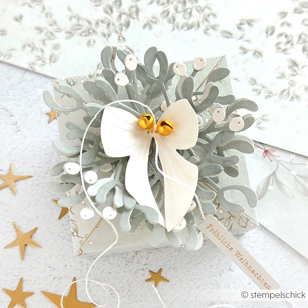 Eine festliche Alexandra Renke Geschenkbox, verziert mit weißem Band, goldenen Glöckchen, silbernem Laub, gefertigt aus Designpapieren und der Motivstanze Mistelzweig Box, umgeben von funkelnden Goldsternen.