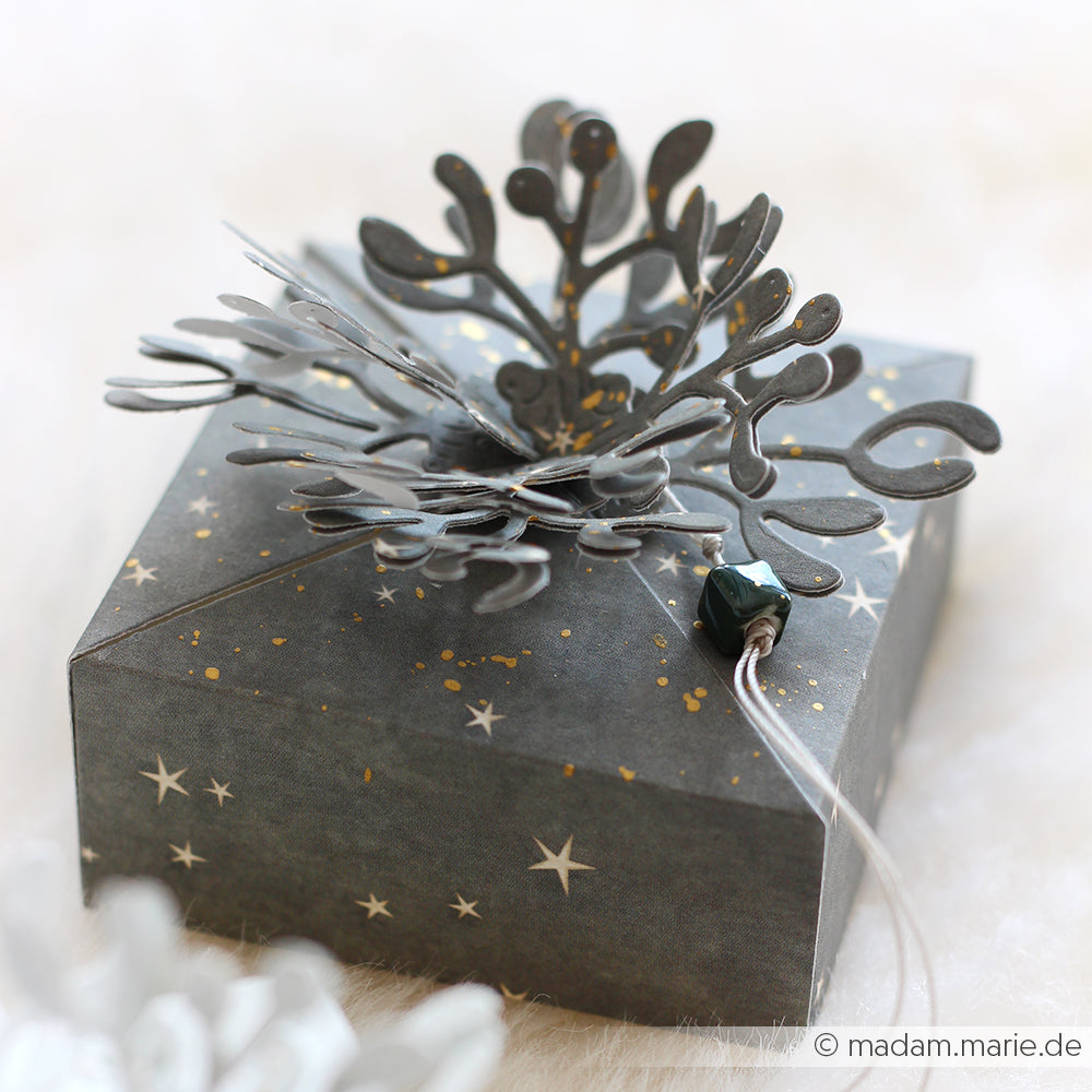 Eine dunkle Alexandra Renke Geschenkbox mit weißen Sternen, dekoriert mit DieCut Systemen, veredelt mit einem silbermetallischen Blumenornament und einer schwarzen Perle.
