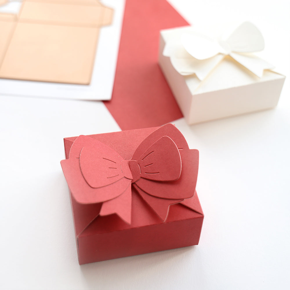 Zwei Geschenkboxen mit großen Papierschleifen - eine rot, eine weiß - gebastelt mit Alexandra Renke Motivstanze Schleifenbox, platziert auf einer rot-weißen Fläche.