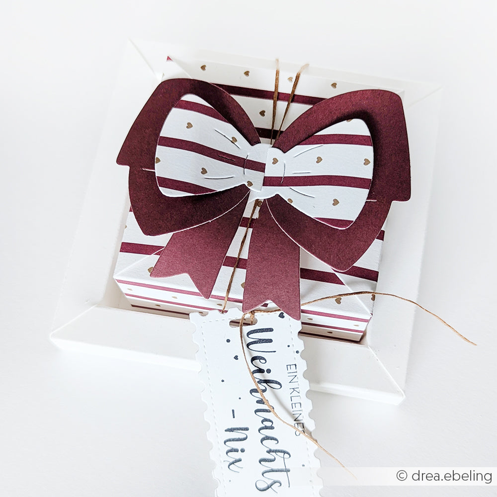 Ein Geschenkkarton mit einer großen weinroten Schleife, gestreiftem Alexandra Renke Geschenkpapier und einem festlichen Anhänger steht auf einer weißen Fläche.