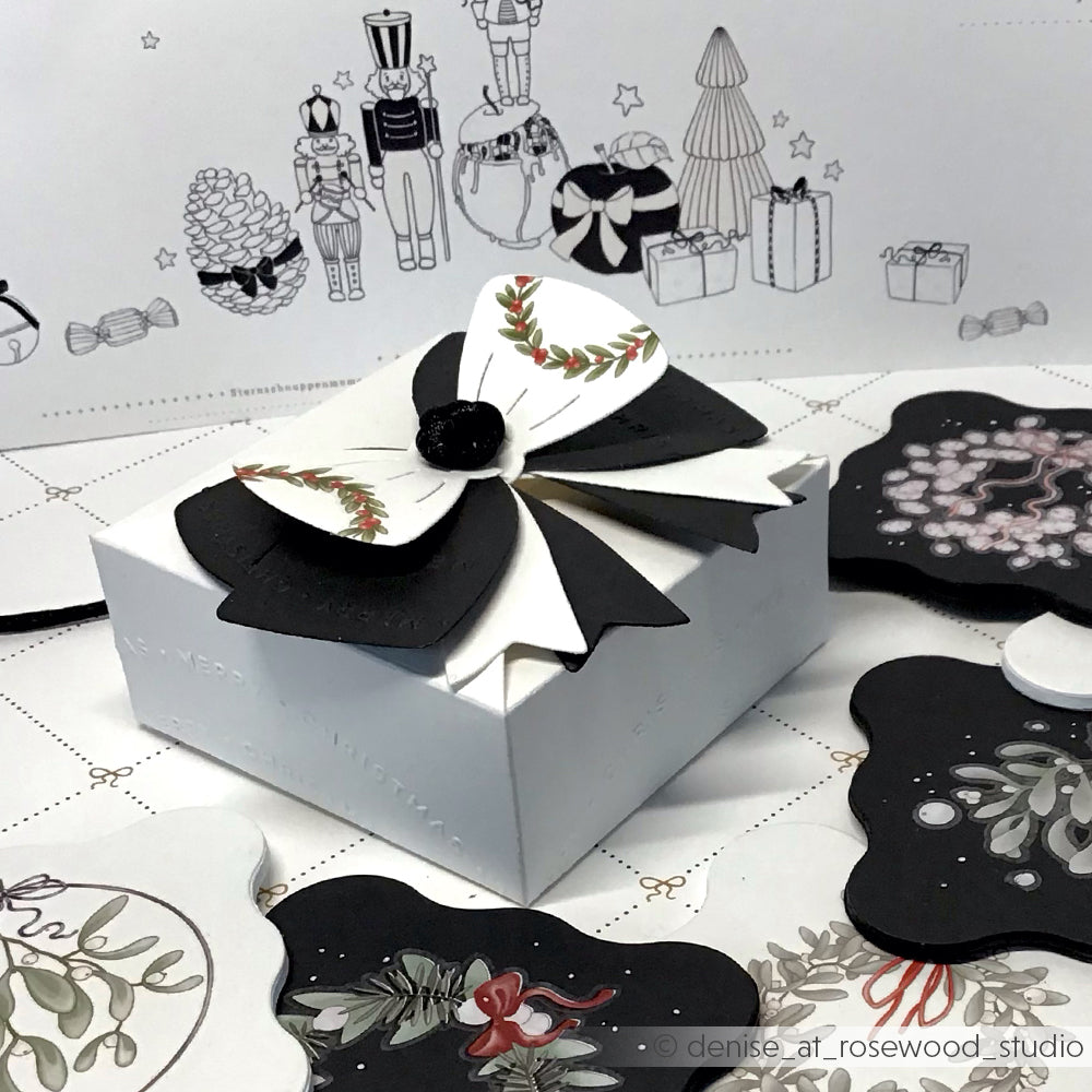 Eine weiße Geschenkbox mit einer schwarz-weißen Schleife, umgeben von weihnachtlichen Karten und Zeichnungen, unterstreicht Alexandra Renkes kreative Designpapiere und die Eleganz der Motivstanze Schleifenbox.