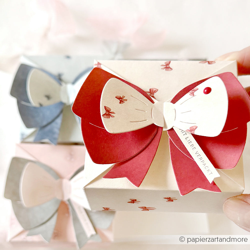 Eine Hand hält eine dekorative Geschenkbox mit einer großen rot-weißen Schleife, roten Schmetterlingsmustern und aufwendigen Details, die mit Alexandra Renke Motivstanze Schleifenbox und eleganten Designpapieren gefertigt wurde.