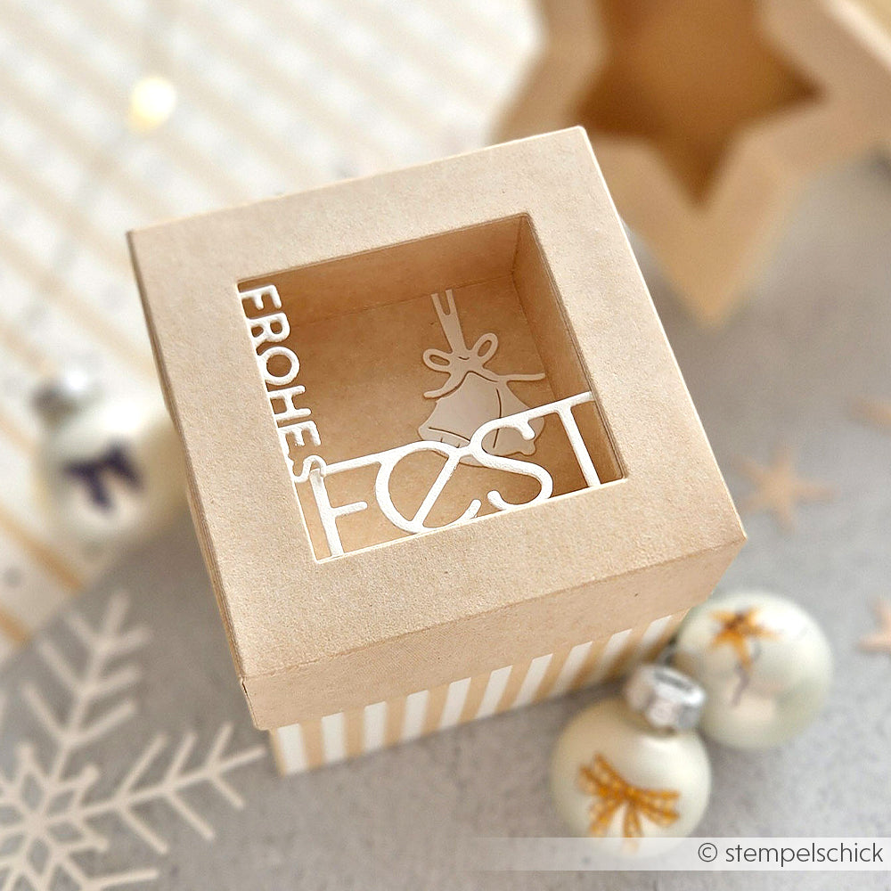 Quadratische Geschenkbox mit Fensterdeckel und der Aufschrift "Frohes Fest" sowie einer Glocke, hergestellt mit den Alexandra Renke Motivstanze quadratische Box Dies. Weiße Ornamente im Hintergrund sorgen für einen festlichen Touch.