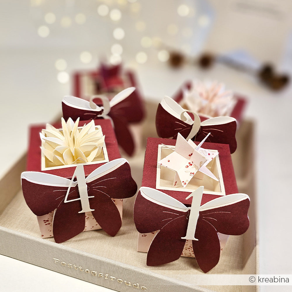 Vier dekorative Geschenkboxen aus der Alexandra Renke Motivstanze Quadratische Box Stanzen mit burgunderroten Schleifen und Zahlen. Präsentiert auf einem Tablett mit weichem Hintergrund unterstreichen sie die Eleganz der DieCut Systeme für besondere Anlässe.
