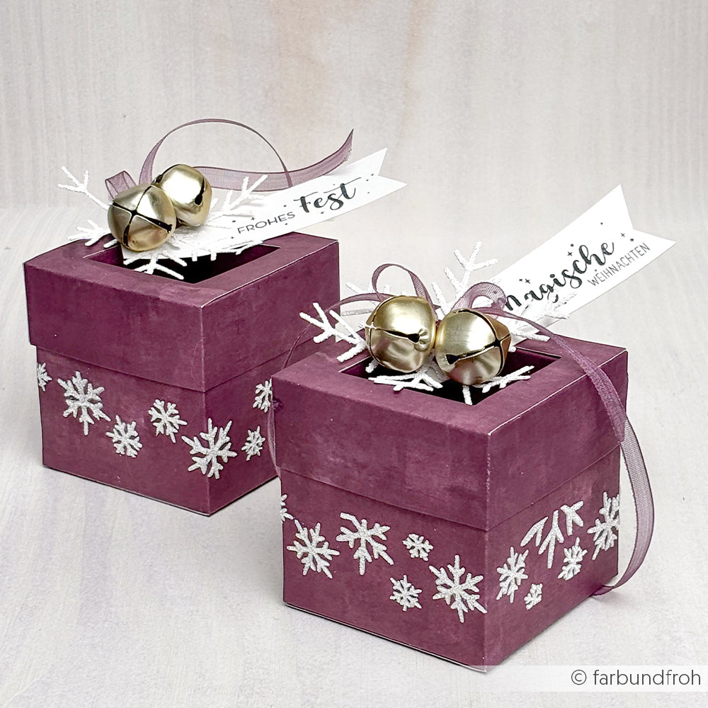Zwei lilafarbene Geschenkschachteln mit weißen Schneeflocken und goldenen Glöckchen sind mit Bändern und festlichen Anhängern verziert und sehen dank Alexandra Renke Designpapieren, die mit einer Motivstanze Quadratische Box gefertigt wurden, elegant aus.