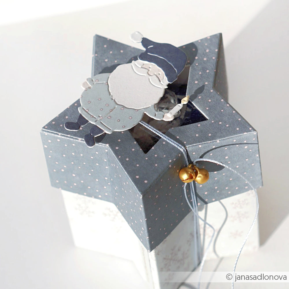 Sternförmige Geschenkbox aus der Motivstanze Stern-Box von Alexandra Renke, verziert mit einem blauen Weihnachtsmann und goldenen Glöckchen, die mit einer Schnur an der Spitze zusammengebunden werden.