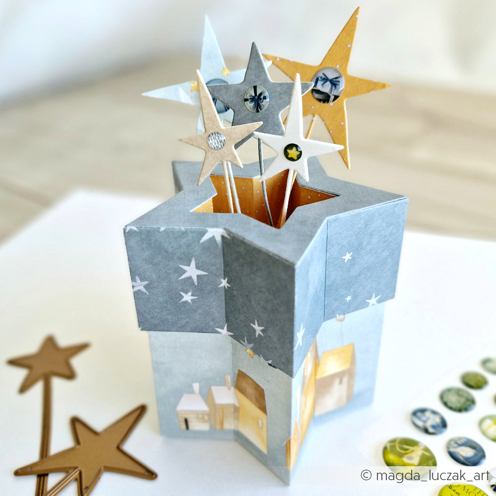 Eine sternförmige Pop-up-Karte von Alexandra Renke, die aus Designpapier gefertigt und mit Sternen und kleinen Häusermotiven verziert ist, liegt auf einem Tisch, der von Sternenausschnitten umgeben ist.