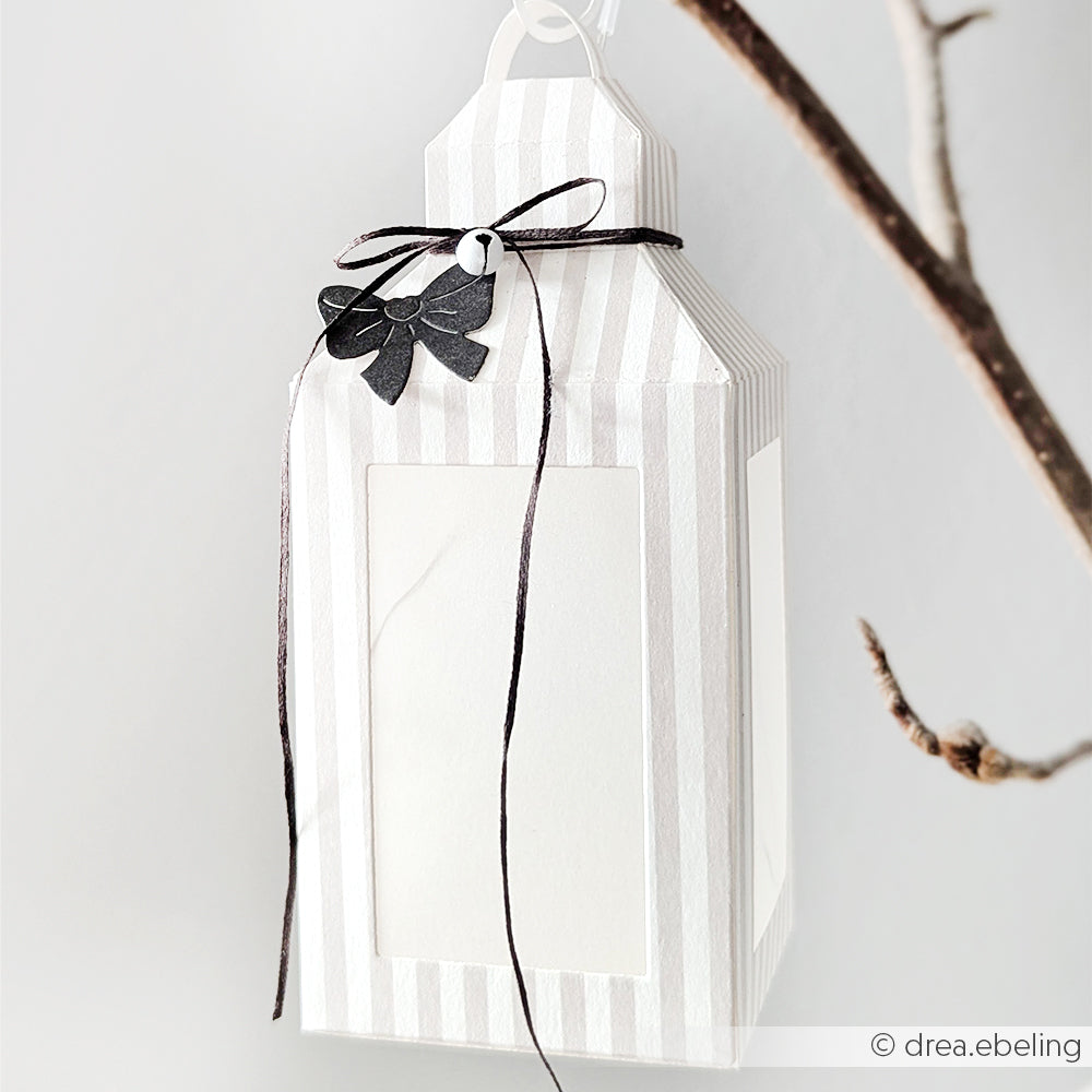 Gestreifte weiße Papierlaterne mit schwarzer Schleife und Blattdekoration, gefertigt aus eleganten Alexandra Renke Designpapieren, die an einem Ast hängen.